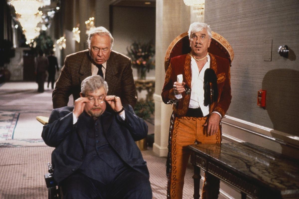 A scene from “Naked Gun 2 1/2: The Smell of Fear,” with George Kennedy (standing) playing Nielsen’s police supervisor and Richard Griffiths (seated) playing a presidential energy adviser. (MovieStillsDB)