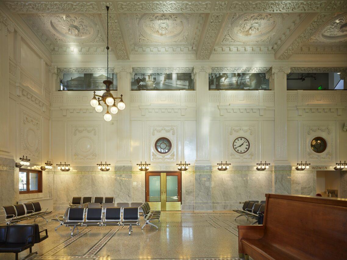Symmetry and sumptuousness define King Street Station’s waiting area. The highly ornamental plaster ceilings and inset fluted Corinthian columns draw up the eye, as do the bronze globe chandelier and wall sconces. The terrazzo floor has inlaid square mosaic tiles. Contrasting the opulence are the simple, round porthole windows flanking the large clock. Black and white historic photographs are featured where the ceiling and wall meets. (Courtesy Photographer Benjamin Benschneider)