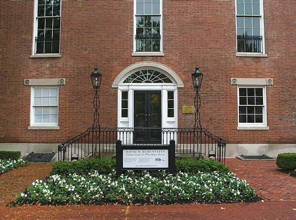 Rosettes carved in stone above windows flank Decatur House’s entrance. Over the entryway’s French lited (clear glass) doors with sidelights is a fanlight pediment window topped with a band of carved stone molding. A wrought iron railing in front of the double-approach is topped with two decorative wrought iron columns and lanterns, adding to the symmetry of the façade’s design. Framing the entryway garden and stairs is a red-brick walkway laid in a basketweave pattern. (Photography by Bruce White)