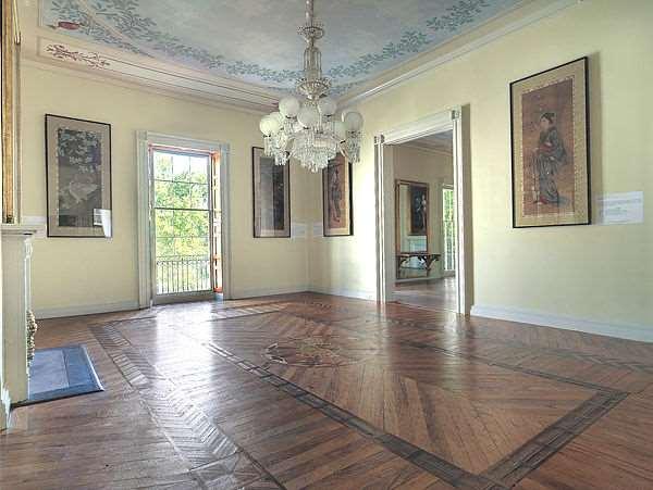 An elaborate, geometric parquet flooring design contrasts with a flowery blue-sky ceiling painting in the formal dining room. The 12-armed chandelier’s style is baroque, similar to lighting found in France’s Palace of Versailles. Frosted globes and multifaceted glass prisms form the chandelier. Accessible from the room’s windows is a narrow Juliet balcony. (Photography by Bruce White)