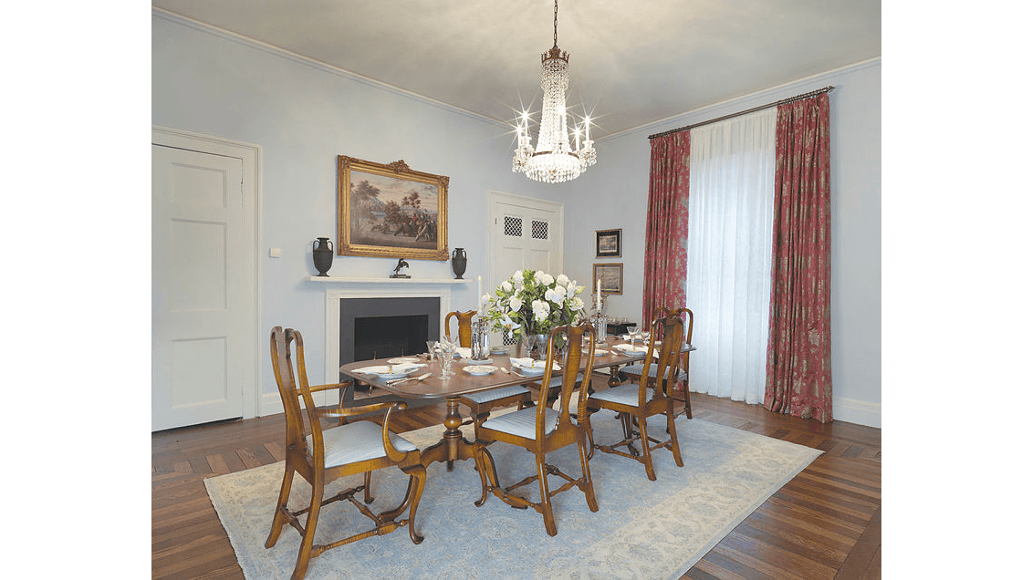 The informal family dining room is apportioned with a reproduction 19th-century table set with antique china and silverware. The simple mantel is offset by a two-tone stained parquet floor and a crystal chandelier. The saturated gold and wine hues on the walls and window treatments coordinate with the 1867 gilt-framed oil-on-canvas painting “Horses Quenching Their Thirst, Camels Disdaining,” by artist Erneste Etienne de Francheville Narjot, which hangs over the mantel. (Photography by Bruce White)