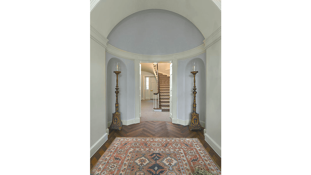 Matching faux vault niches for housing floor lighting flank the front hall doorway, over which is a shallow dome, in contrast to a full hemispherical (or half-circle) dome. Over the dome is an archway accentuated by wide crown moldings. A chevron-design (stripes that meet in a “V” pattern) parquet floor peeks out from beneath an English-made Wilton carpet.  (Photography by Bruce White)