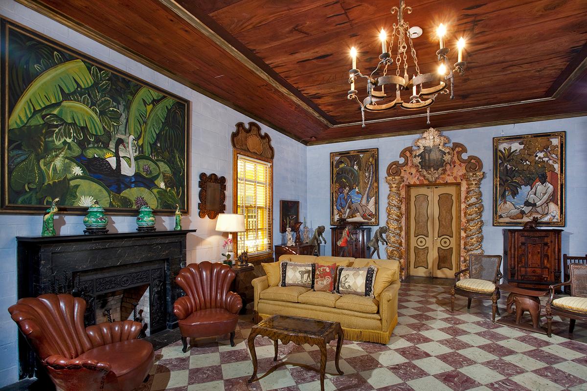 The house’s drawing room is replete with eclecticism. Italian baroque-style Solomonic columns, with their distinctive spiral shape, flank the door; gilded and gray-painted carvings of acanthus leaves wind around the columns, which are topped with decorative Corinthian capitals. The door and decorative window plaques were painted by Bartlett to resemble terrazzo and marble. The tray ceiling includes a mahogany center that is surrounded by local Dade County Pine. The red and white stone, which makes up the tiled checkerboard floor, was quarried in Florida. Queen Anne leather fan back chairs and a tasseled-fringe gold sofa are the focal point furnishings situated at the solid black marble fireplace. The eight-light, wrought-iron chandelier is Spanish baroque in style. (Courtesy of Bonnet House)