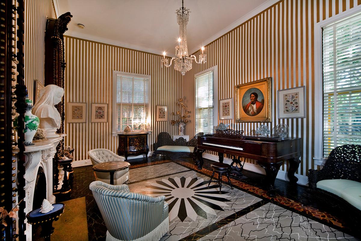 In the music room is an 1872 Steinway piano made of rosewood, with ivory keys. Bartlett painted the floor to resemble marble and terrazzo, with a large black-and-white flower design as the centerpiece. The rest of this glamorous space, with gold-and-white wallpaper, button-tufted and fringed upholstery, and a crystal chandelier, is conveyed in the decor's blend of art deco and Victorian styles. Prominent in the room is a replica of the bust "Veiled Woman" by Italian sculptor Giovanni Battista Lombardi (1823–1880). (Courtesy of Bonnet House)