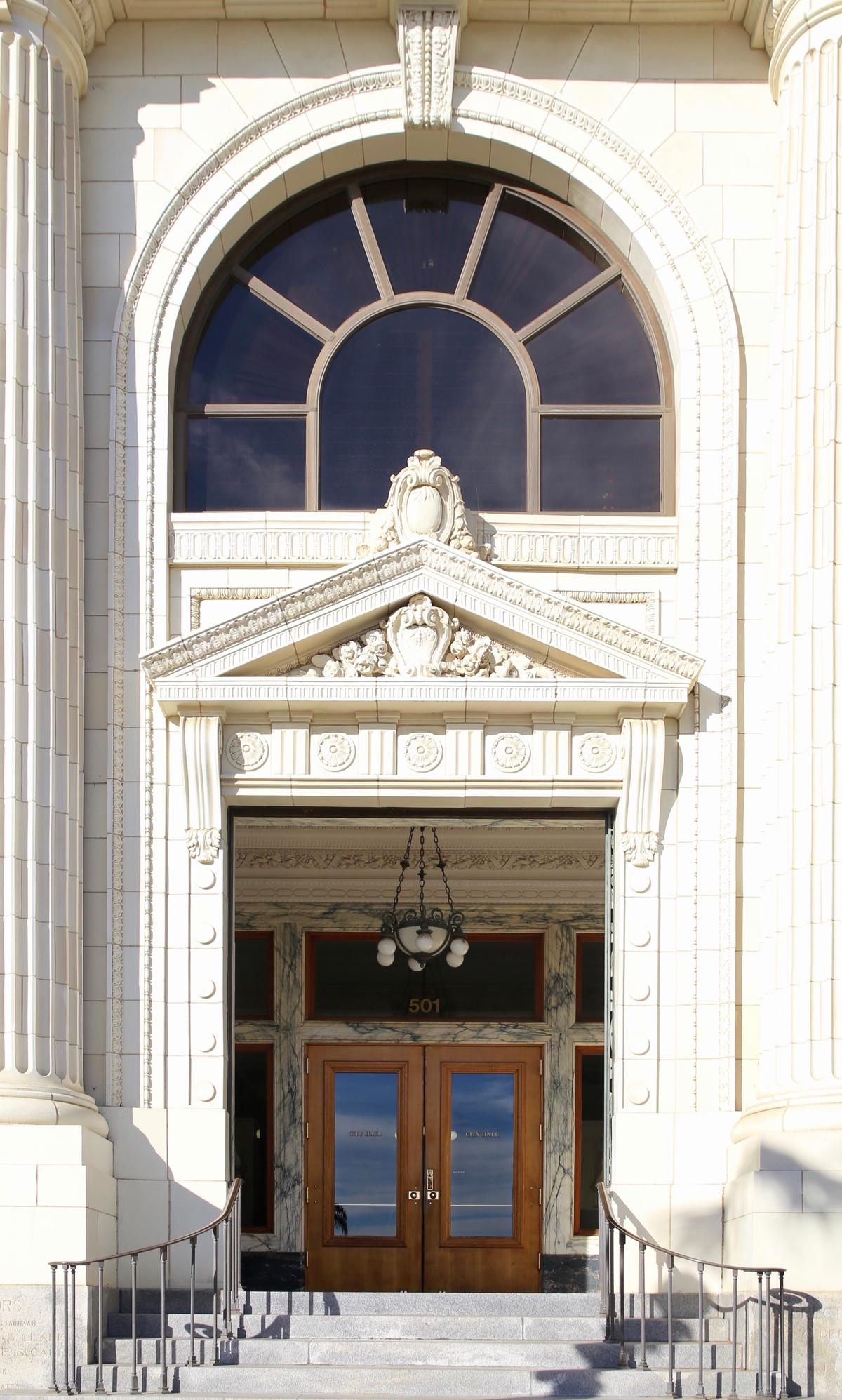 Black and gold calacatta marble, from Carrara, Italy, outlines the windows and wooden double doors, which are inset into an open vestibule. A brass, dome-light chandelier hangs in the center of the entryway. The portal of the granite stairway is heavily bedecked with carvings of columns, rosettes, acanthus leaves, roping, and cartouches. (City of Ventura)