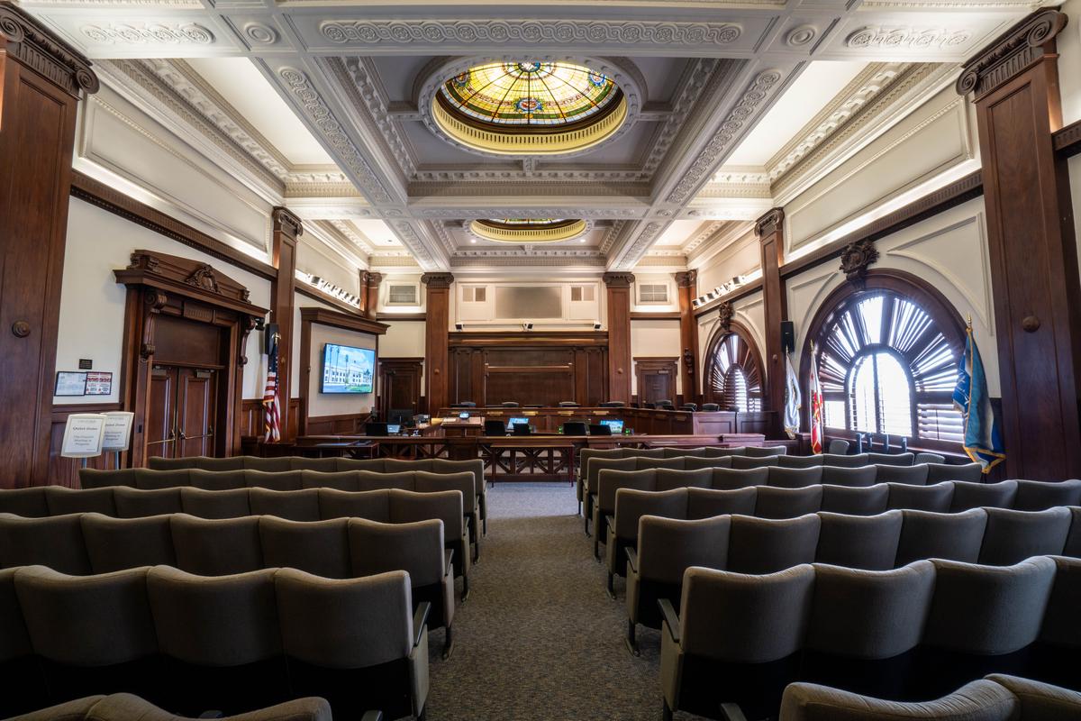 The interior council chamber on the second floor was previously used as a courtroom. A tray ceiling inset with stained glass skylights has the appearance of being supported by numerous carved mahogany columns topped with Ionic capitals. Carved mahogany woodwork surrounds the doorway and arched windows and is used for the railing, dais (where council members sit), and back wall. (City of Ventura)
