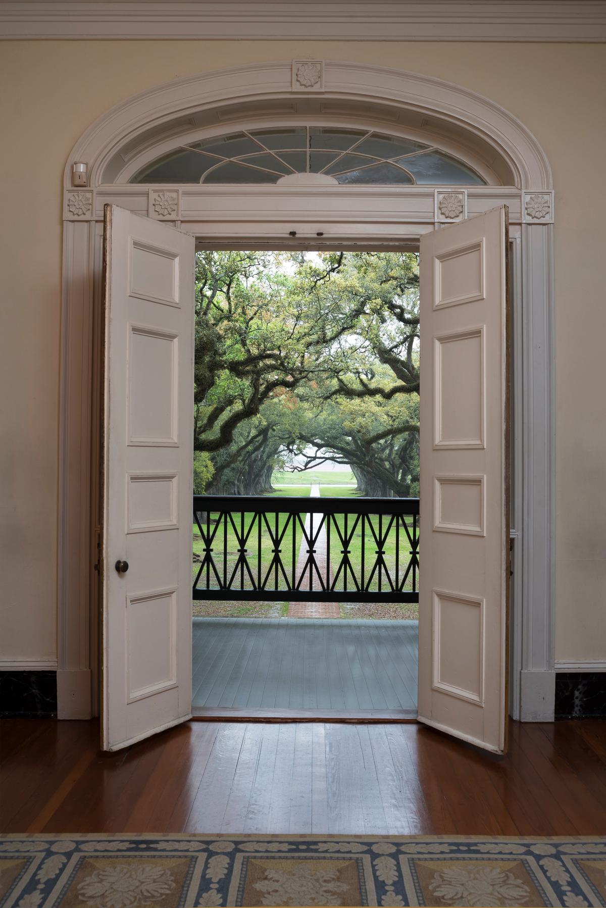 Paneled doors to Oak Alley Plantation’s mansion sit under an arched, sunburst transom featuring a rosette molding in place of a capstone. Rosette moldings also decorate the doorframe. The exterior view from the open door is of the bundled wheat wood railing that runs along the mansion’s upper balconies. (Oak Alley Foundation)