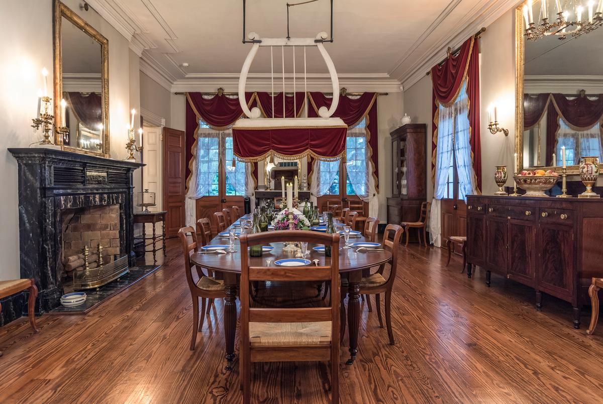 Over the dining room table is what was known as a shoo-fly fan, which is original to the mansion. Resembling the shape of a lyre, the idea of the unique fan originated in East India and was called a “punkah.” The intent is for someone, typically a house slave in antebellum times, to pull on a rope to make the fan swing slowly back and forth while people ate at the dining table. The air flow discouraged flies from landing on food and provided a breeze on sweltering summer days. Crimson velvet drapes with gold-tassel trim hang regally on the tall windows below the molded tray ceiling. (Oak Alley Foundation)