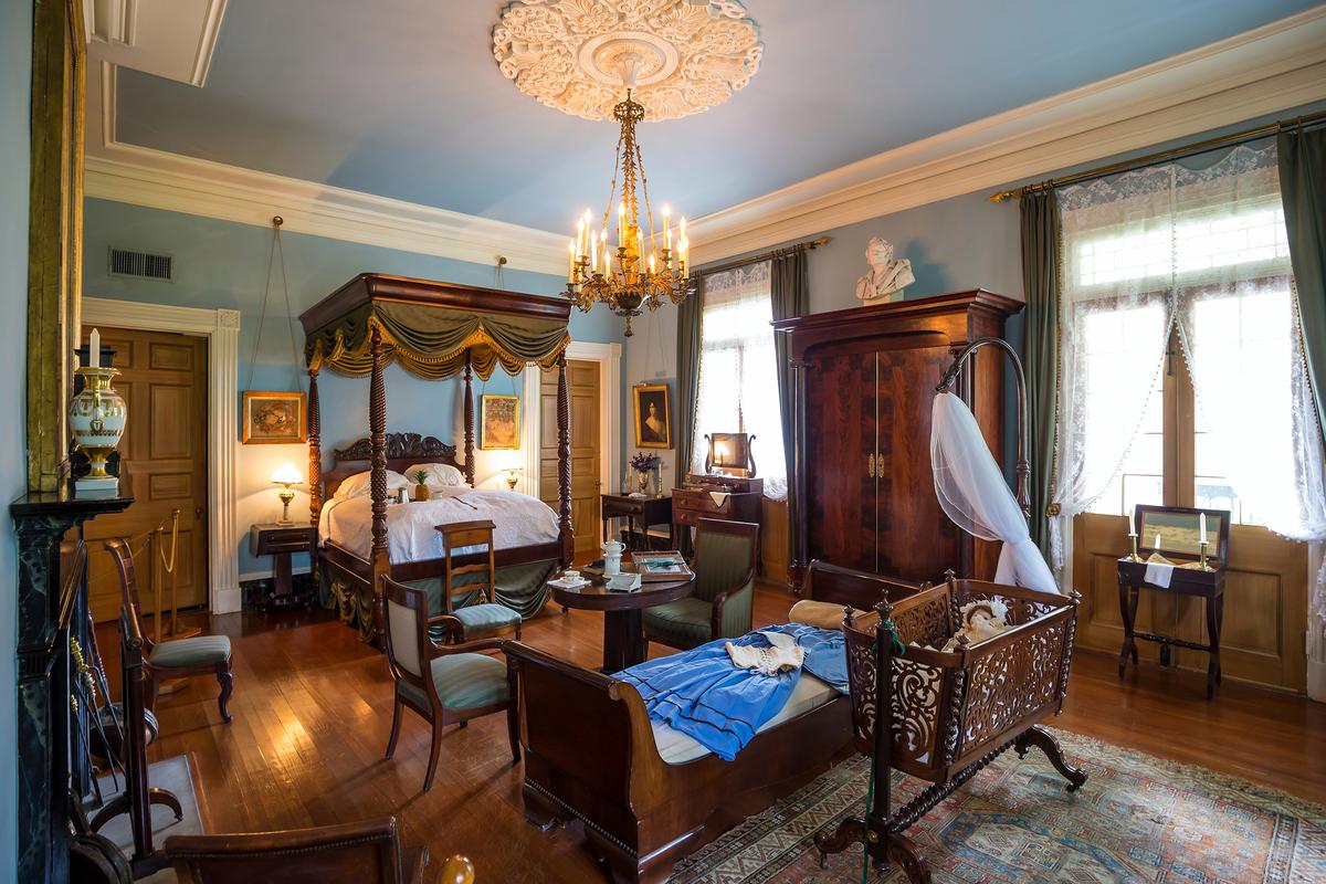 Each of the main rooms at Oak Alley Plantation exhibit an exceptional and distinct ceiling medallion, from which hangs an equally singular lighting fixture. This upper-floor bedroom is no exception. Densely furnished with handcrafted 19th-century period antiques, centerpiece items in this room are the sleigh-style daybed and the intricately carved cradle on a turned base with Chippendale legs. The carved four-poster, canopy bed is outfitted with forest-green velvet with gold-tassel trim that hangs from the bed’s tester, or top frame. (f11photo/Shutterstock)