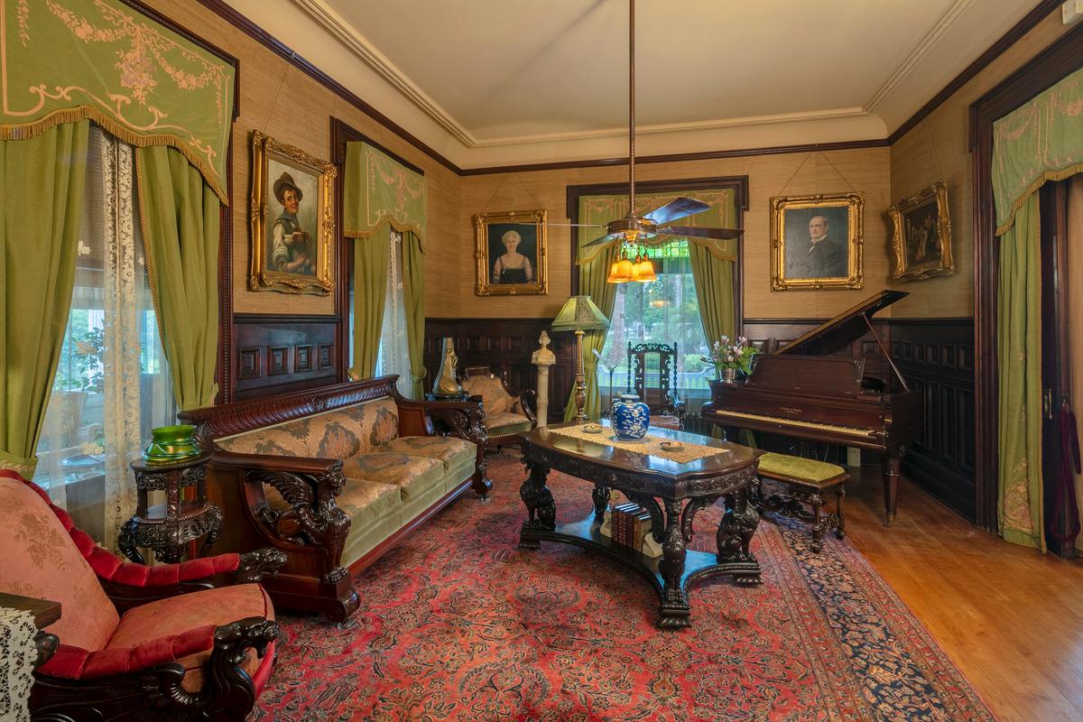 The mansion’s music room features a mahogany grand piano, manufactured in Boston. Reproduction green-hued, tasseled window draperies sport lyre images intertwined in a floral garland; hanging from the fabric cornice are lace curtains. A high, carved birchwood chair railing gives way to grasscloth on the walls. Hanging from the tray ceiling are gilt-framed portrait paintings. (McFaddin-Ward House)