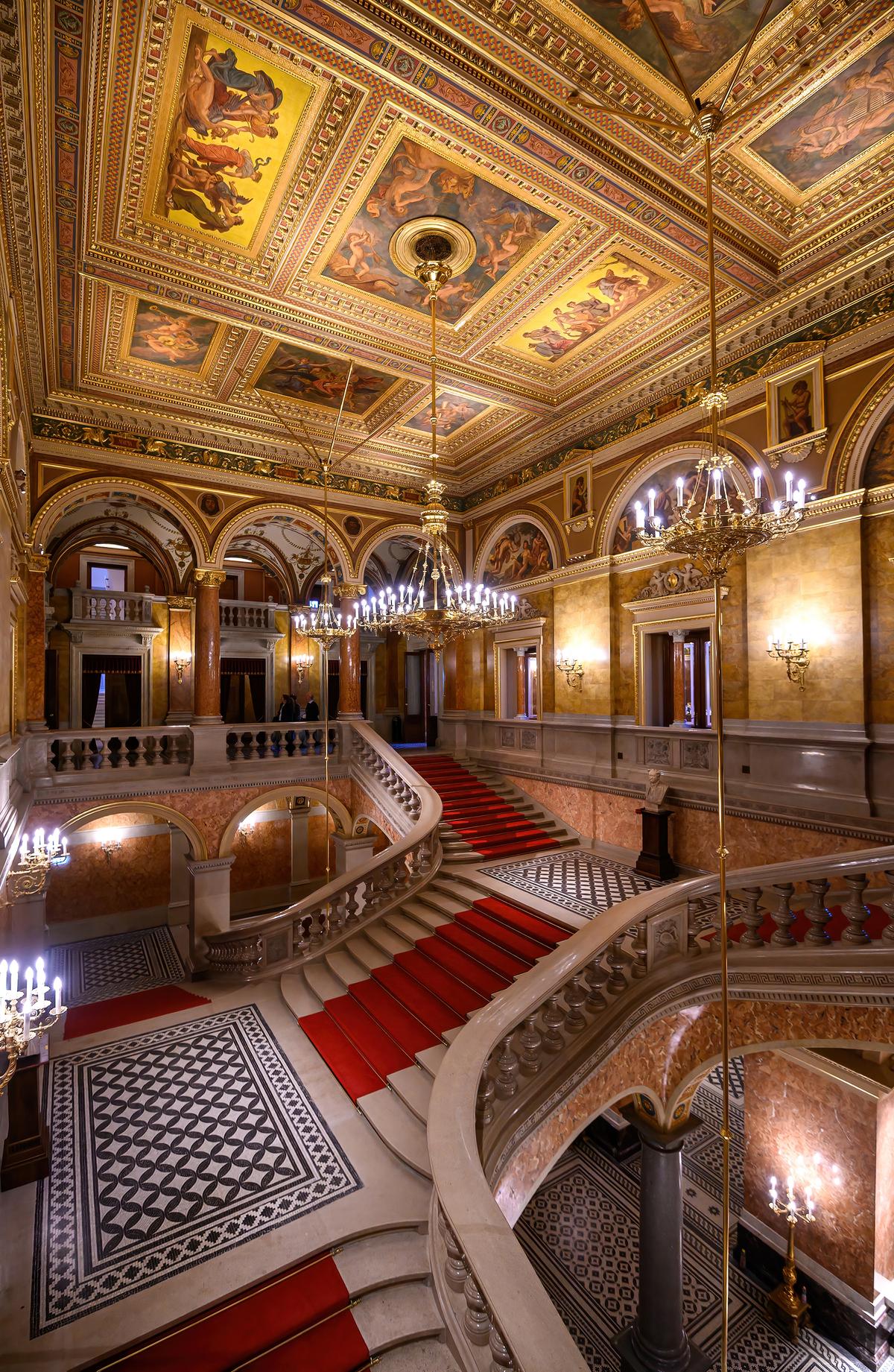 Sweeping in two directions inside the Hungarian State Opera House is the bifurcated foyer staircase leading to what is considered the upper-level Grand Staircase. Crimson-red carpet runners over the marble stairs provide visiting patrons with a truly regal experience as they approach auditorium entrances. The landing is made of patterned tiles. The aesthetics of this space are greatly enhanced by the mythological, music-themed paintings in the nine rectangles composing the gilded, coffered ceiling. (Mitzo/Shutterstock)