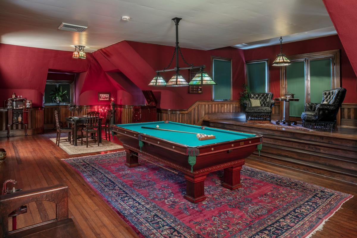 On the spacious third floor is a billiard room with a masculine ambiance expressed in the button-tufted leather chairs on the platform and the dark-stained wood furnishings. The Arts and Crafts-style, stained glass fixture over the chairs, billiard table, and card table provide some color to the room. The walls are covered in a burgundy burlap. Wood types in the room are oak, poplar, and maple. (McFaddin-Ward House)
