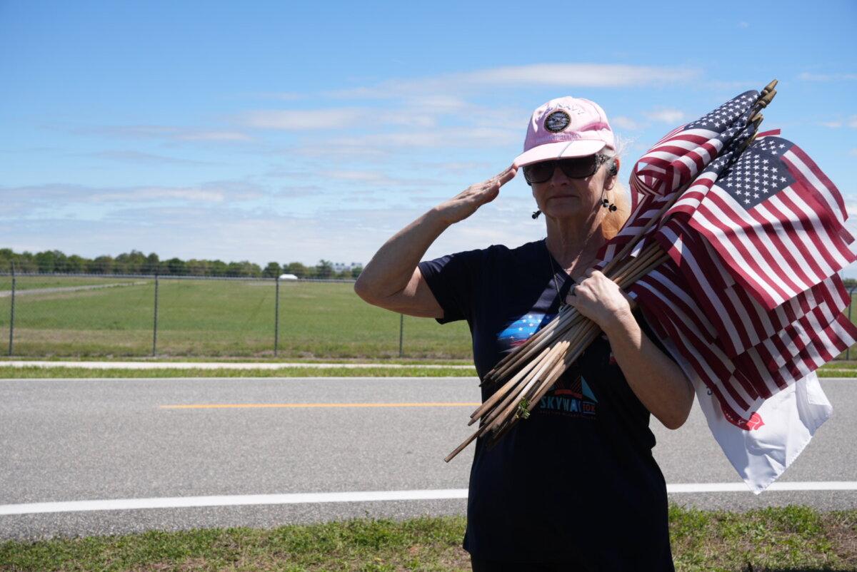 Laura Dantin, a U.S. Navy veteran, stood at attention when the funeral procession for one of the fallen U.S. soldiers from the Iran War drove by her. She helped place out some of the hundreds of American flags that lined the roadways in Winter Haven, Fla., on March 18, 2026. (Troy Myers/The Epoch Times)