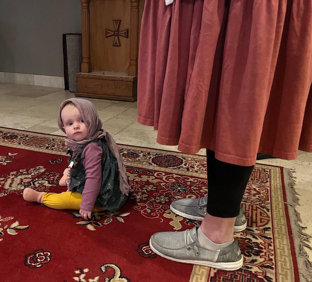 A little girl in traditional Russian attire sits at her mother's feet during a service at Holy Archangels Orthodox Church in Phoenix on Jan. 30, 2026. (Allan Stein/The Epoch Times)