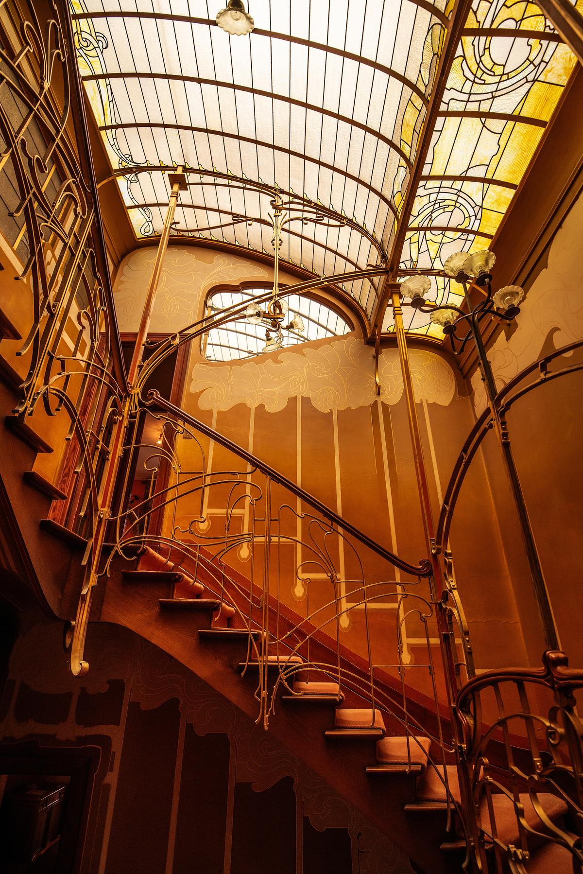 Above the main staircase, a large curved stained-glass skylight with golden tones floods the space, highlighting the warmth of the interiors. This innovative design transforms the house’s central core, traditionally dark and difficult to use, into a bright, inviting area that serves as both a functional circulation space and a striking focal point of Horta’s art nouveau vision. (Veggiewayfarer/Shutterstock)