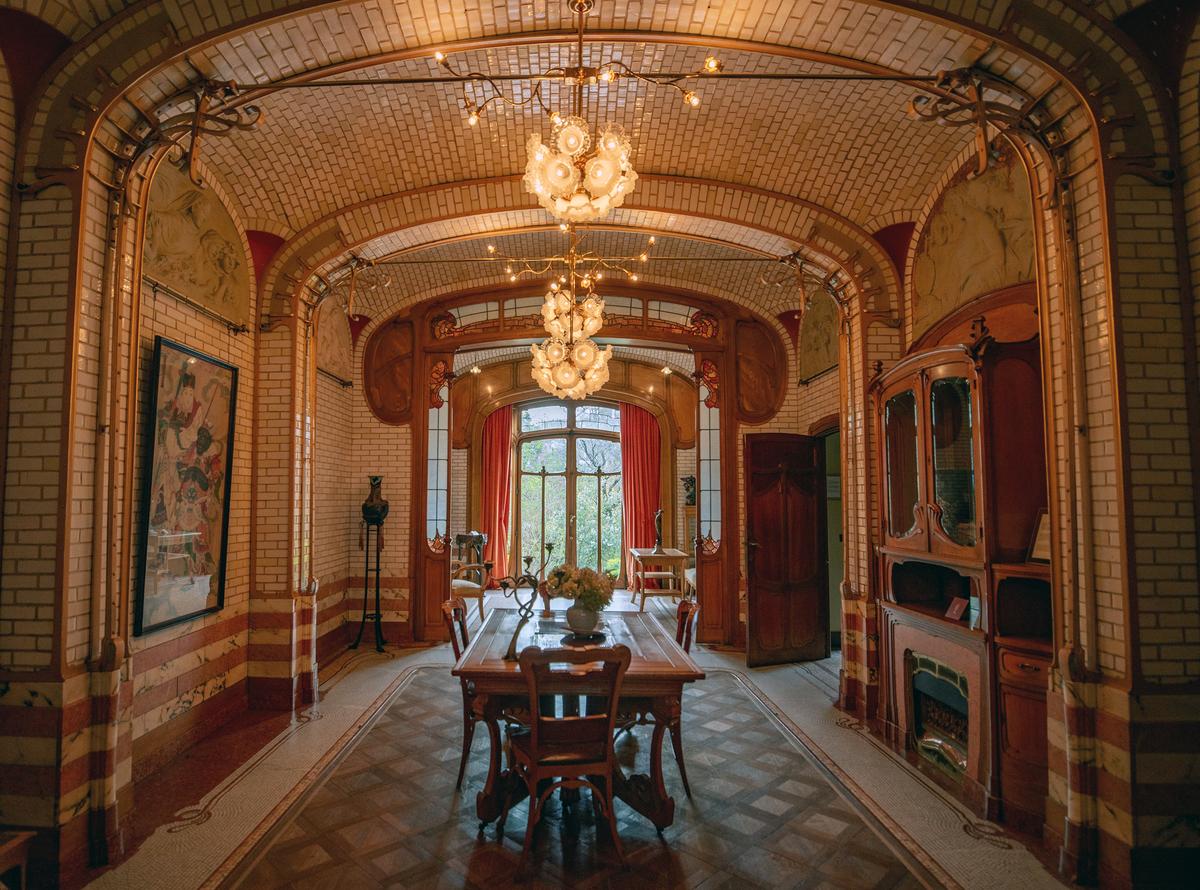 The dining room features a meticulous design of white glazed bricks, exposed metal arches, and parquet flooring bordered by a tile mosaic. Ornate, overhanging light fixtures complement the large doors that open to the garden, filling the space with both natural and decorative light. (Veggiewayfarer/Shutterstock)