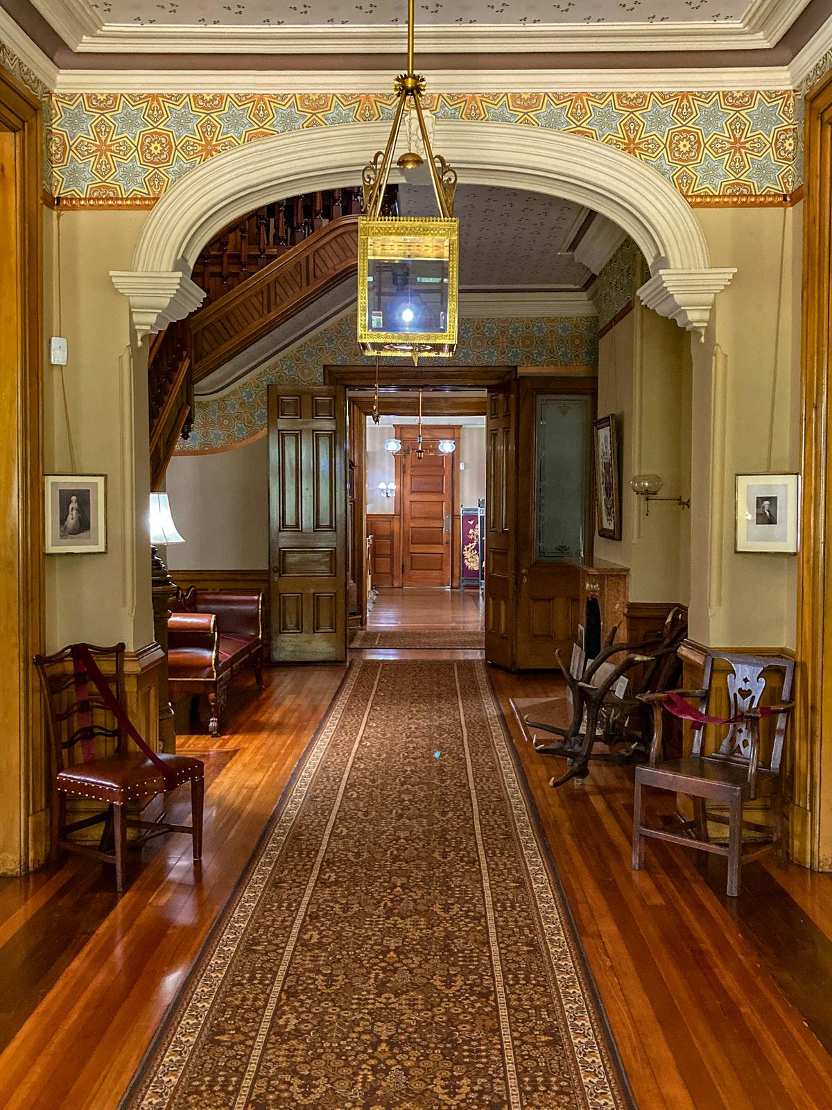 The entrance hall greets guests with a Victorian display of 19th-century wallpaper and a half-arch with a keystone and stacked molding cornices. The florid brass and etched glass gasolier is one of the many distinct lighting fixtures found hanging prominently in each room. Tucked to the left of the hallway is a cantilevered, four-level staircase. (Hayes Presidential Library & Museums)