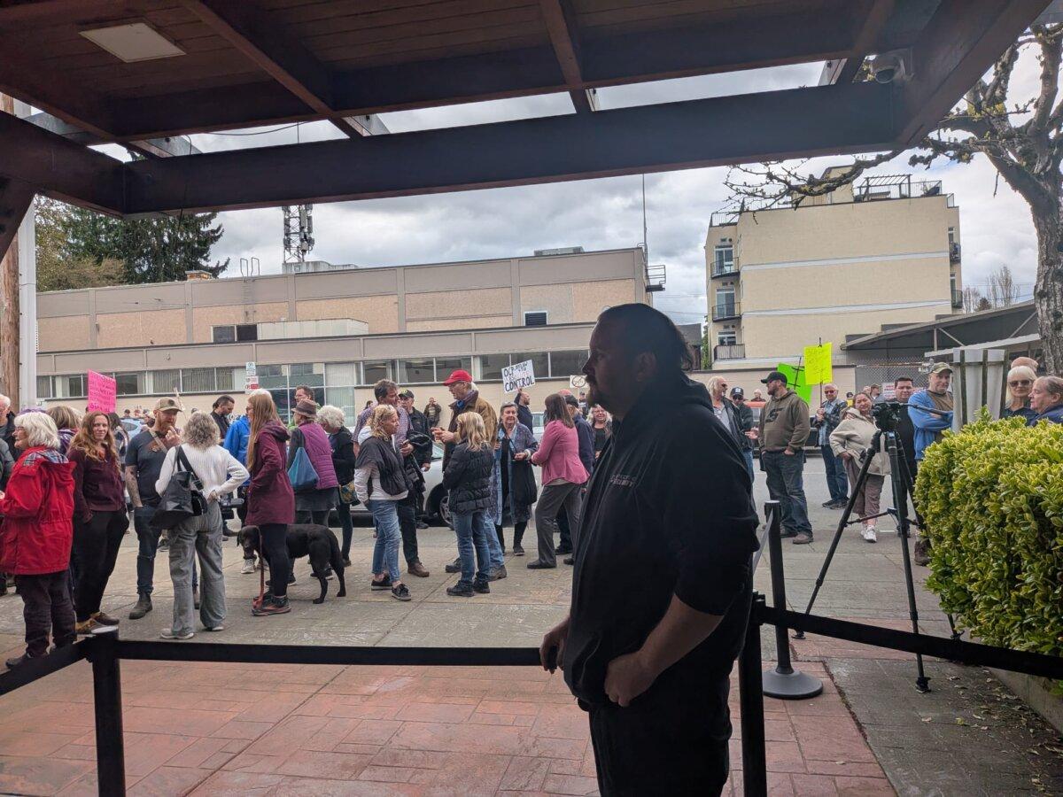 Cowichan Valley residents protest outside the Cowichan Valley Regional District office in Duncan, B.C., as a security guard stands by on April 22, 2026. (Paul Rowan Brian/The Epoch Times)