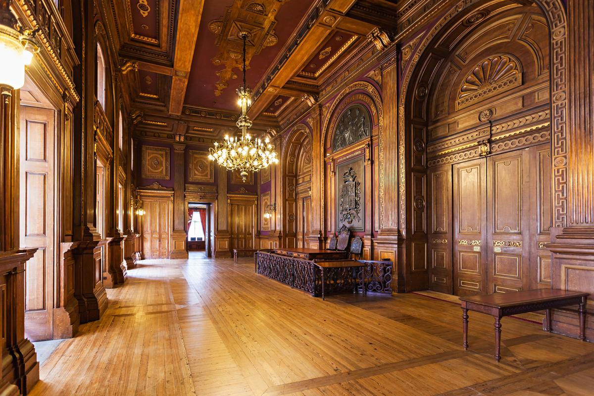 On the third floor is the Assembly Room. While each room in this grand structure is distinct, this room may be the most different due to its ambiance created by floor-to-ceiling, carved wood. However, much of what appears to be wood, such as doors, ceiling, and walls, is actually painted plaster that gives the appearance of wood. The wooden flooring<span style="color: #ff0000;"> </span>exhibits a simple parquet pattern. The room’s adornments are the paintings in the tray ceiling and the centerpiece chandelier, which weighs approximately 1 ton. (saiko3p/Shutterstock)