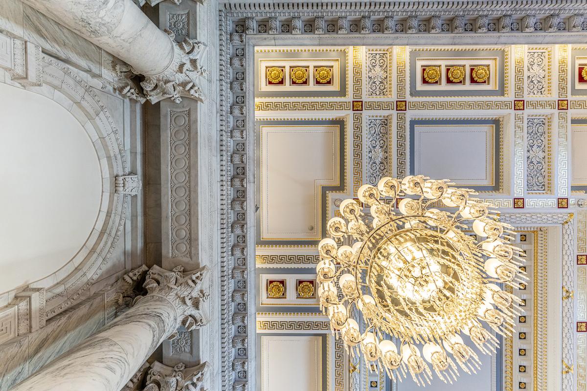 While there is so much to admire in the Kennedy Caucus Room, its ceiling is a key architectural achievement. The elegant coffered ceiling is bordered with a frame of carved acanthus leaves. The design is outfitted with such telltale beaux arts designs, such as gilded rosettes and a band of the classic, interlocking Greek key pattern. Some of the ceiling spaces are trimmed in blue to coordinate with the gilded accents. (Public Domain)