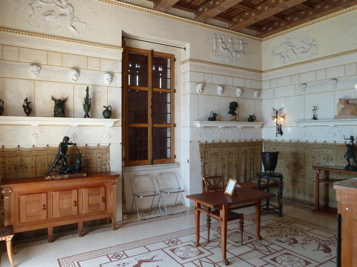 The “oikos” is a room dedicated to the arts. Its stucco frieze of bas-relief carvings showcases musical instruments and a range of exaggerated theater masks, a key component of ancient Greek performances. A lemonwood cabinet (peeking out from the bottom right)<span style="color: #ff0000;"> </span>conceals a miniature folding piano. While not accurate for the period, it was a request from Reinach’s wife. (<a href="https://commons.wikimedia.org/wiki/File:Villa_kerylos_oikos.JPG" target="_blank" rel="nofollow noopener">Abxbay</a>/ <a title="Category:CC-BY-SA-3.0" href="https://commons.wikimedia.org/wiki/Category:CC-BY-SA-3.0">CC-BY-SA-3.0</a>)