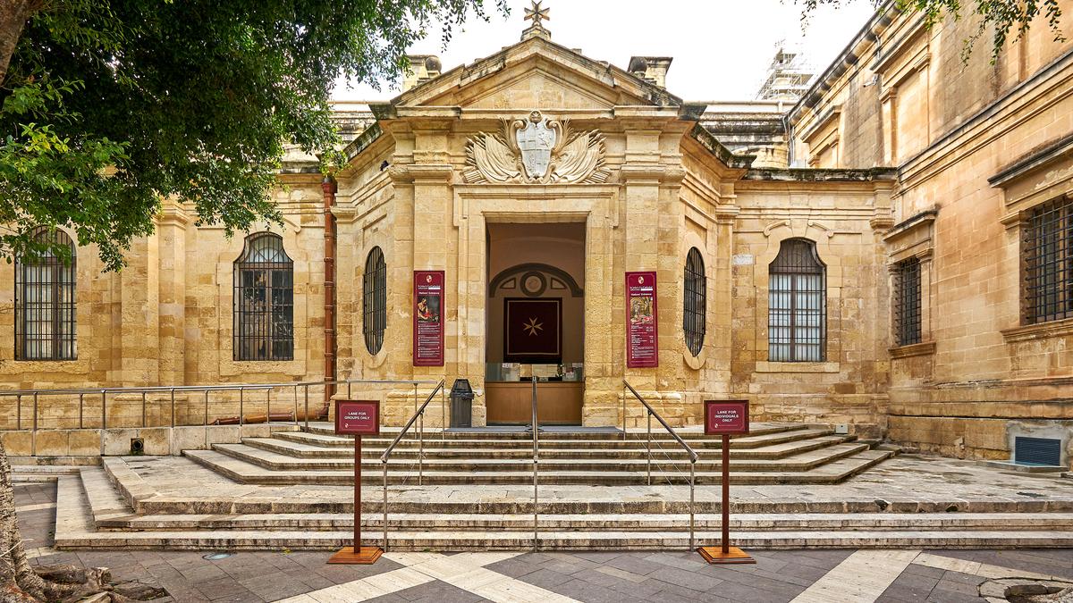 The main entrance of St. John’s Co-Cathedral is set within a Maltese limestone façade, approached by a short stairway and framed in smooth ashlar with restrained sculptural detail. It is surmounted by the heraldic escutcheon of the Order of St. John, marking its status as a conventual church.  (Vladimir Zhoga/Shutterstock)