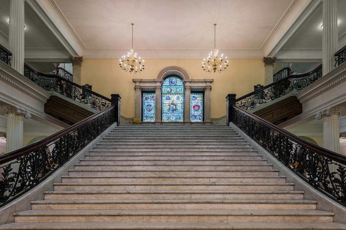 The grand staircase features elegant granite treads on the horizontal surfaces of each step, paired with wrought-iron railings. A balustrade of carved wood and detailed ironwork guides movement between floors. A sizable Palladian stained-glass window at the half landing displays Massachusetts seals in vivid color, tracing themes from colonial charters through revolutionary and state government heritage. (Nagel Photography/Shutterstock)