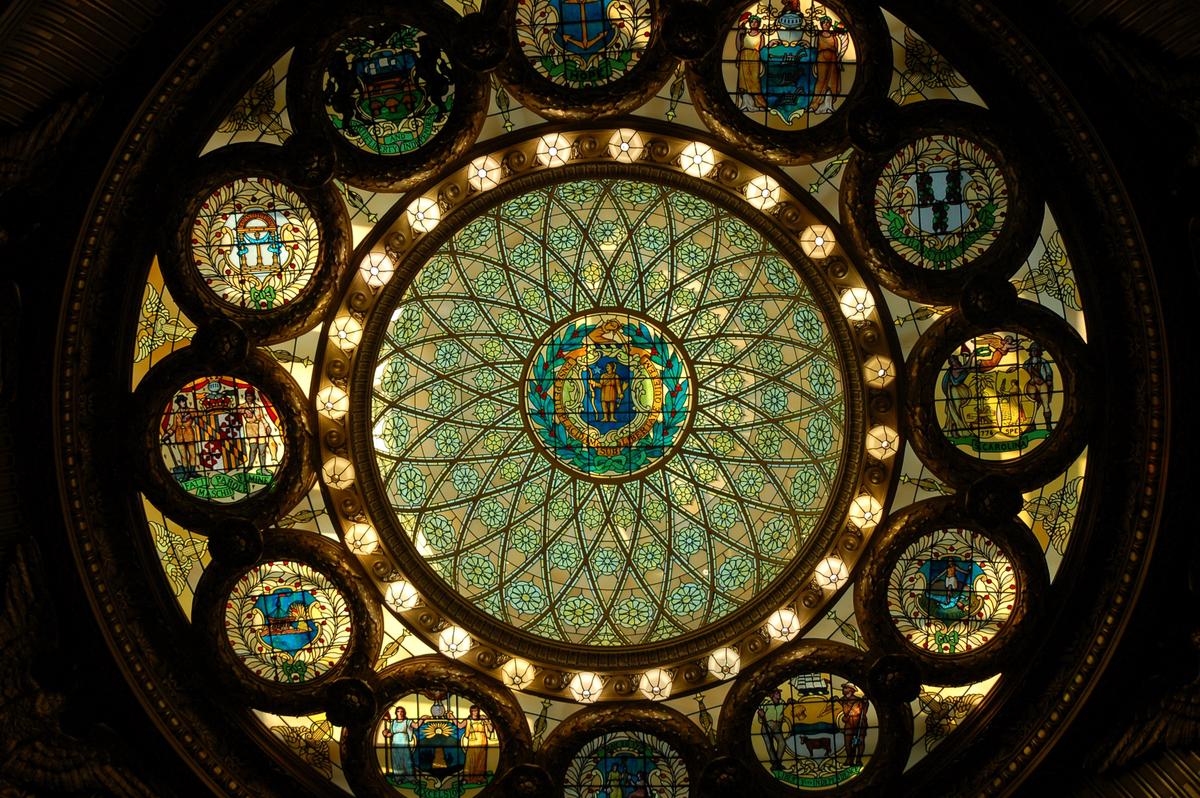 The stained-glass skylight above Memorial Hall features a vivid circular design. At its center is an early Massachusetts state seal depicting an Algonquian Native American with a bow and arrow, surrounded by the seals of the other 12 original colonies. Crafted by Ford and Brooks and installed in 1900, it bathes the Hall in softly filtered light. (James Kirkikis/Shutterstock)