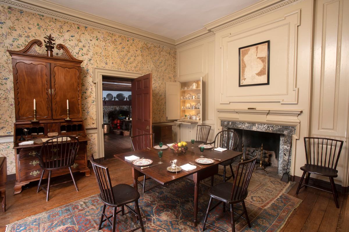 Colonial-era Windsor chairs are situated around the simple dining table; a Windsor chair is also pulled up to the Chippendale-style secretary desk. The ceiling is bordered by crown molding featuring a rope-trim design. A built-in corner hutch holds dishes and china. The walls are paneled, and an overmantel with cross-cut corners frames the marble fireplace. (Courtesy of Historic Odessa Foundation)