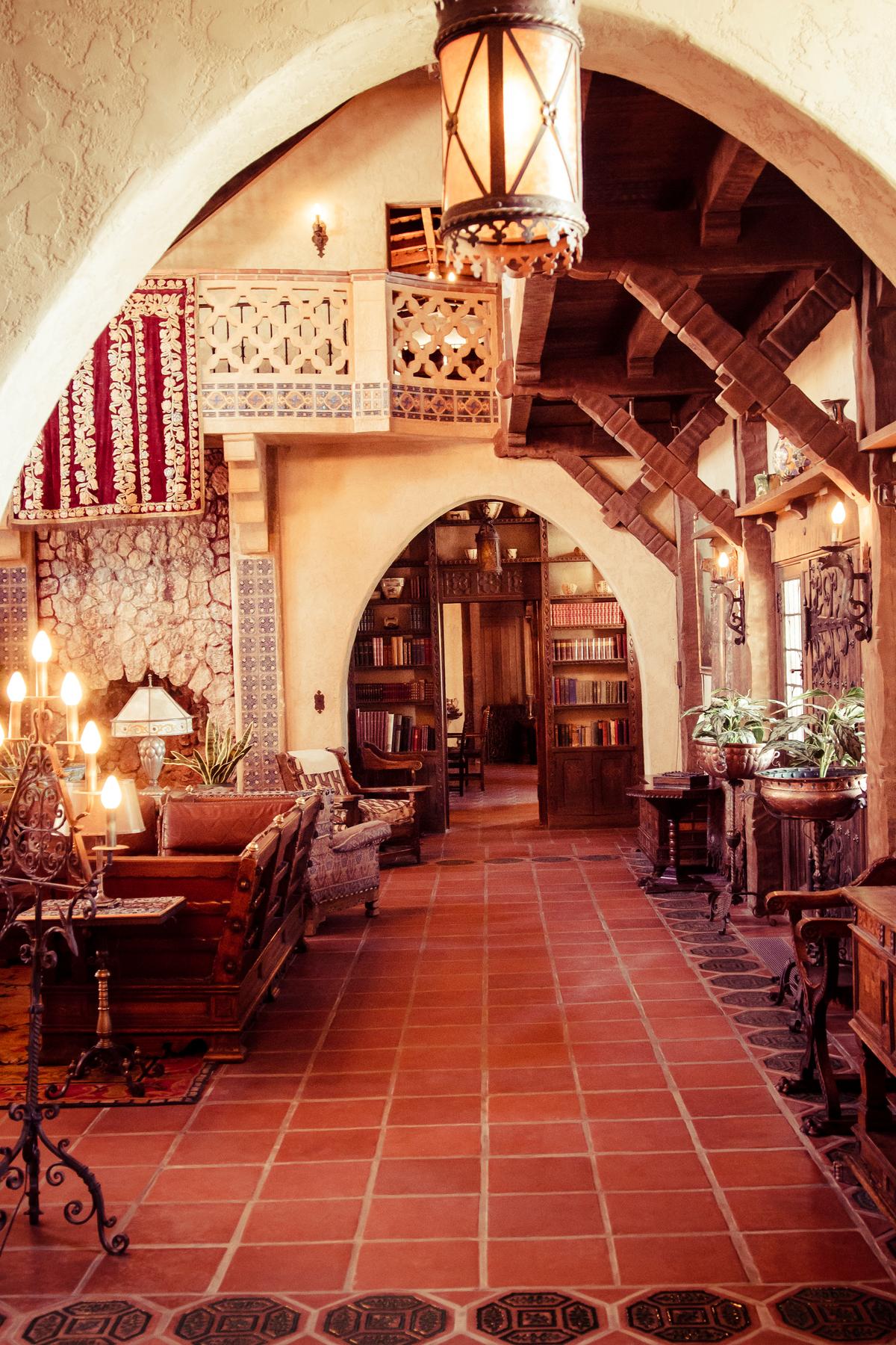 Mexican, Spanish, mission, and Mediterranean influences abound in this lively living room. The quatrefoil-inspired upper balcony and the pilasters flanking the stone fireplace are adorned with painted tiles. Inset in the Gothic arch pass-through, which leads to the dining room, is a built-in bookcase. The rustic knee-brace, red-wood beams are hand-carved. (sprokop/Getty Images)