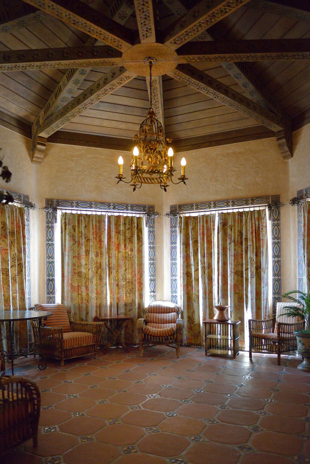With floors of red quarry tile, this sitting room has an octagonal ceiling with exposed beams as its primary adornment. A floral flourished metal candelabra light fixture hangs from a small medallion. Framing the windows is a border of painted tiles. (Cavan-Images/Shutterstock)