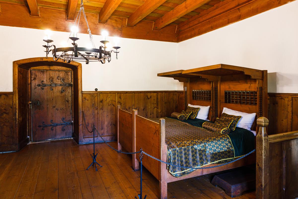 This bedchamber features a half-tester bed with a wooden canopy, dressed in dark green-and-gold patterned coverlets. Dark wood paneling lines the walls beneath a matching wooden ceiling, while a rustic metal chandelier hangs near a heavy arched wooden door, completing the room's sturdy, functional character. (Tartezy/Shutterstock)