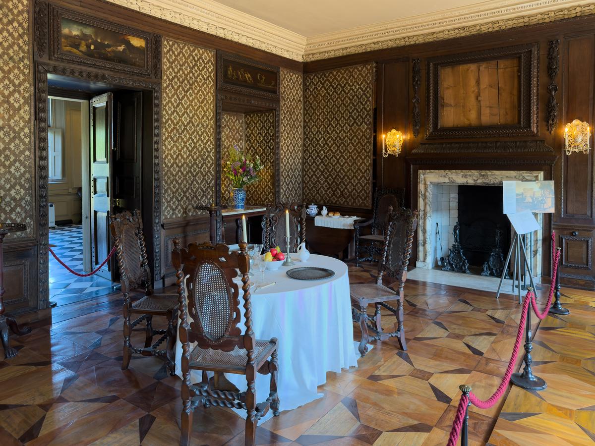 A less ostentatious space at Ham House is the Marble Dining Room. It once had a marble floor; hence, the name. However, during a mid-18th-century renovation, a fine marquetry floor was installed that presents a pattern with tropical hardwoods. The wall coverings are a repeating pattern of four leaves surrounding a rosette. This understated dining space includes a marble fireplace with carved wood paneling, door frames, and doors. (<a title="User:Mike Peel" href="https://commons.wikimedia.org/wiki/File:At_Ham_House_2025_297.jpg">Mike Peel</a>/<a title="Category:CC-BY-SA-4.0" href="https://commons.wikimedia.org/wiki/Category:CC-BY-SA-4.0">CC-BY-SA-4.0</a>)