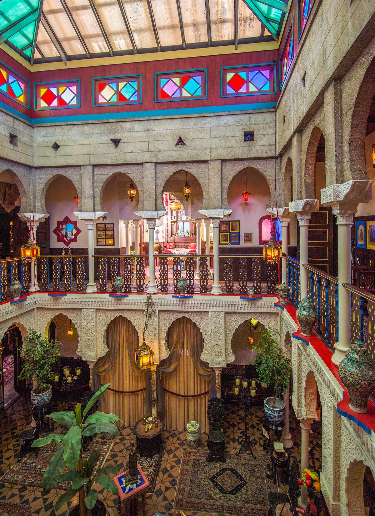 A tented atrium roof allows light to illuminate the vibrant, two-story main hall, dubbed the Court of Lions. A columned balcony with ornate filigree railings surrounds the upper level. Pilasters are between the horseshoe columns, while the entablature features geometric cutouts that complement the geometric stained glass. Fanciful carvings adorn the scalloped arches on the first floor. (Villa Zorayda Museum)