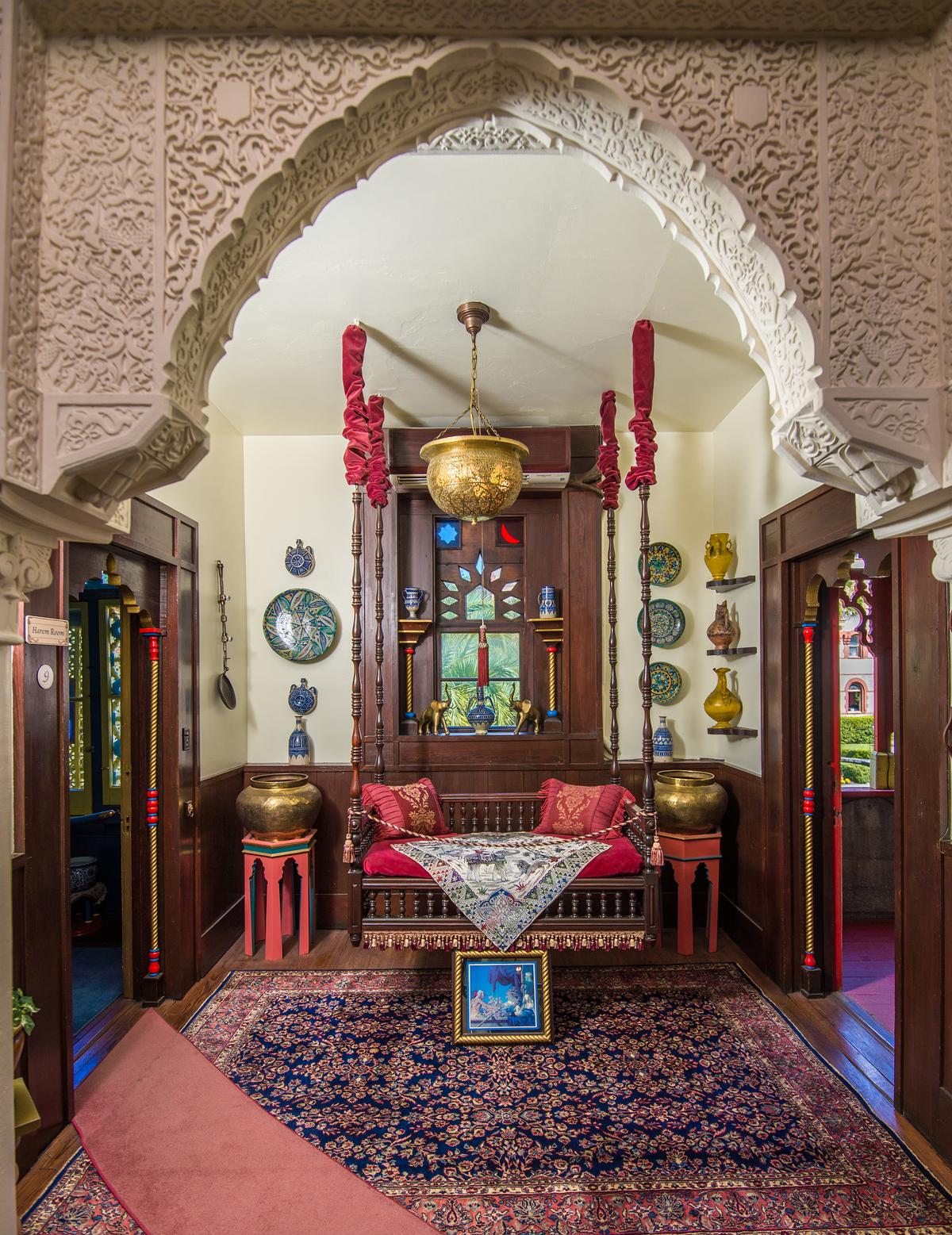 A swinging wooden daybed hanging from long spindles is aligned with this niche space, located next to the harem. Many of the carvings and much of the plasterwork around doors and on walls are exact replicas of those that ornament the Alhambra, a 13th-century palace in Granada, Spain. Every room, including this one, displays oriental rugs, decorative tiles, and fine art collected from Spain, the Middle East, and Egypt. Florida pine, along with imported mahogany and walnut, was used to craft paneling around door frames and at windows. The hand-pierced brass hanging lantern is shaped like the brass bowls flanking the daybed. (Villa Zorayda Museum)