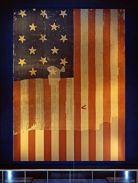 The large Star Spangled Banner Flag that inspired the lyrics of the U.S. national anthem on display at the Smithsonian's National Museum of History and Technology, around 1964. Many pieces were cut off the flag and given away as souvenirs early during its history. A linen backing, attached in 1914, shows the original extent of the flag. (Smithsonian Institution Archives/Public Domain)