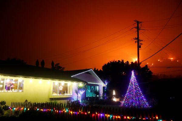 People stand on a roof of a home illuminated with Christmas lights to watch wildfire on a hillside burn during the Thomas Fire in Santa Barbara county near Carpinteria, California, U.S. Dec.11, 2017. (Reuters/Patrick T Fallon)
