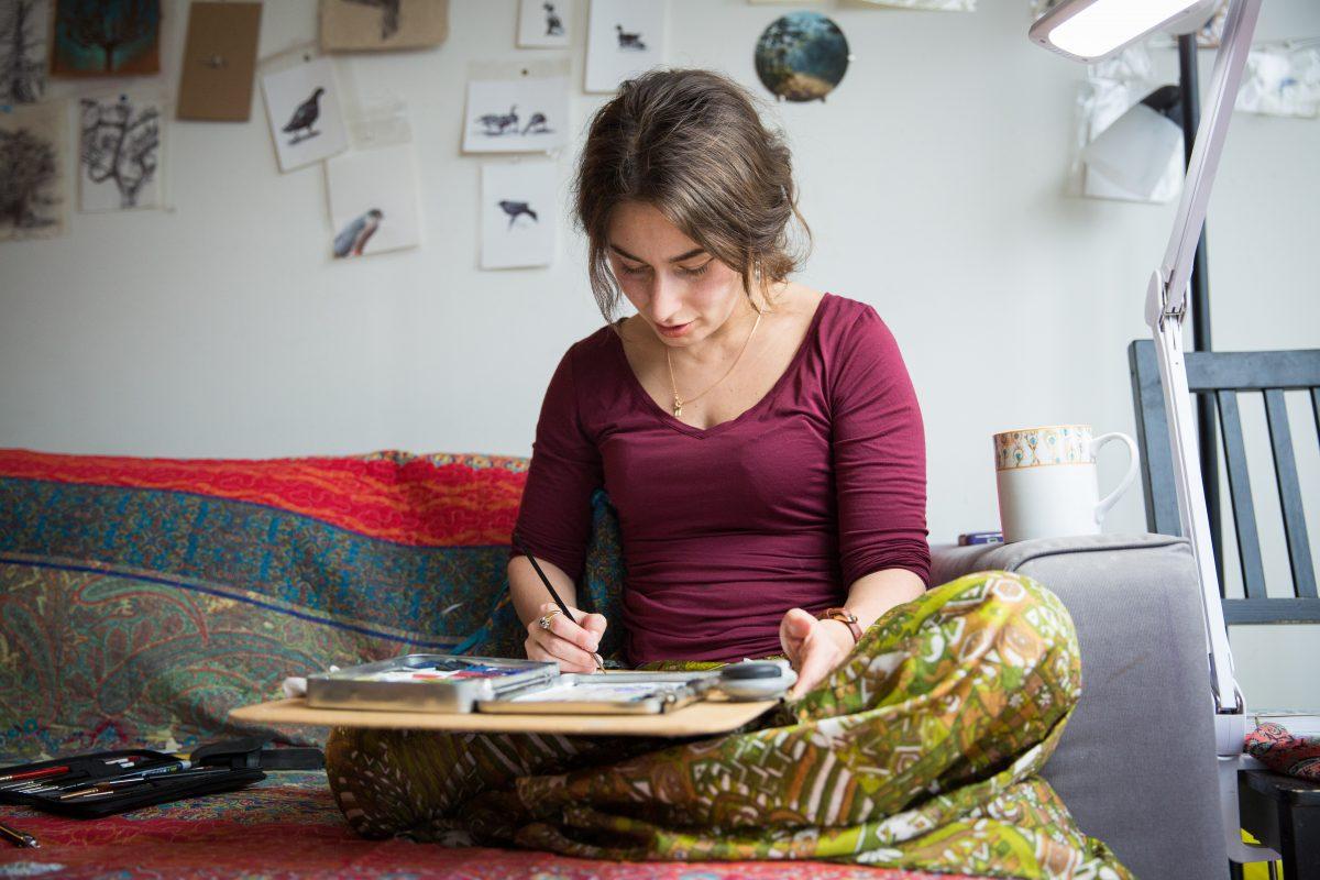 Dina Brodsky paints in the corner of her couch at home in Long Island City in Queens, New York, on Jan. 22, 2018. (Benjamin Chasteen/The Epoch Times)