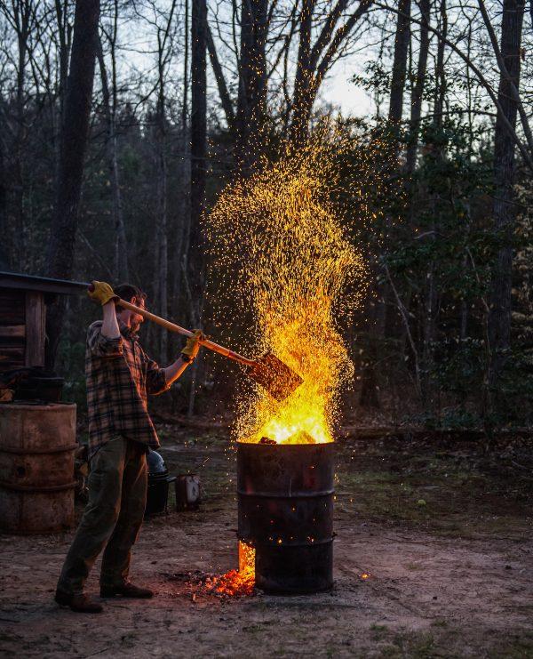 Stone tends to a burn barrel, a centuries-old way of burning fresh wood down to hot coals to use for barbecue. (Ken Goodman​)