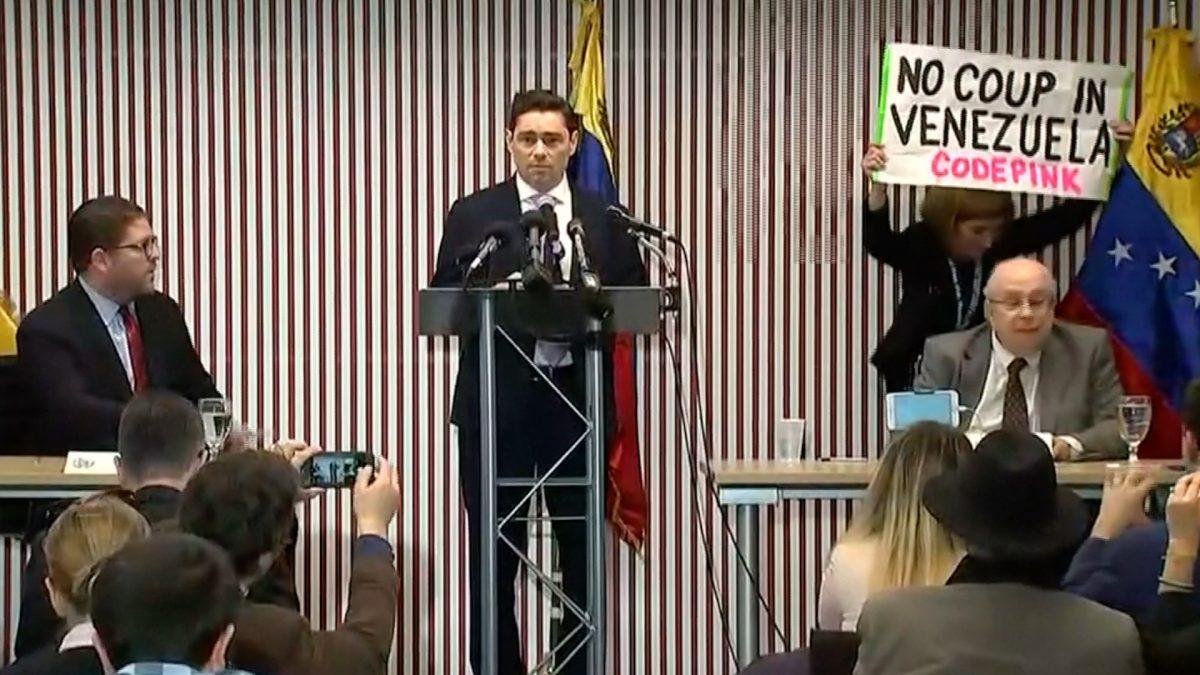 Code Pink protester walks on stage with poster reading 'no coup in Venezuela', while the Venezuelan opposition's envoy to Washington, Vecchio, stops anyone from interfering with protesters, and goes on speaking. Washington, Feb. 21, 2019 (Images via Reuters)