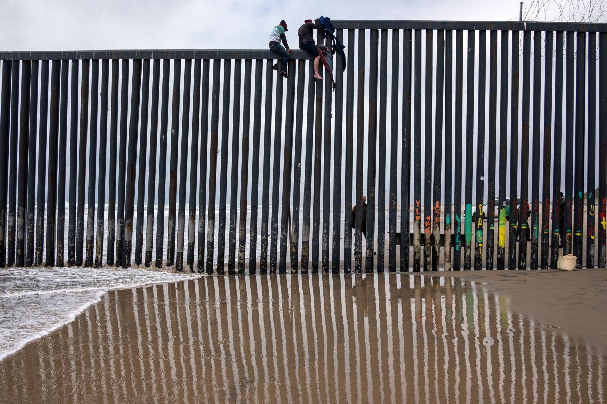 Central American migrants sit above the U.S.–Mexico border fence as a Border Patrol agent stands guard, as seen from Tijuana to San Diego as seen from Playas de Tijuana, Mexico, on March 21, 2019. (Guillermo Arias/AFP/Getty Images)