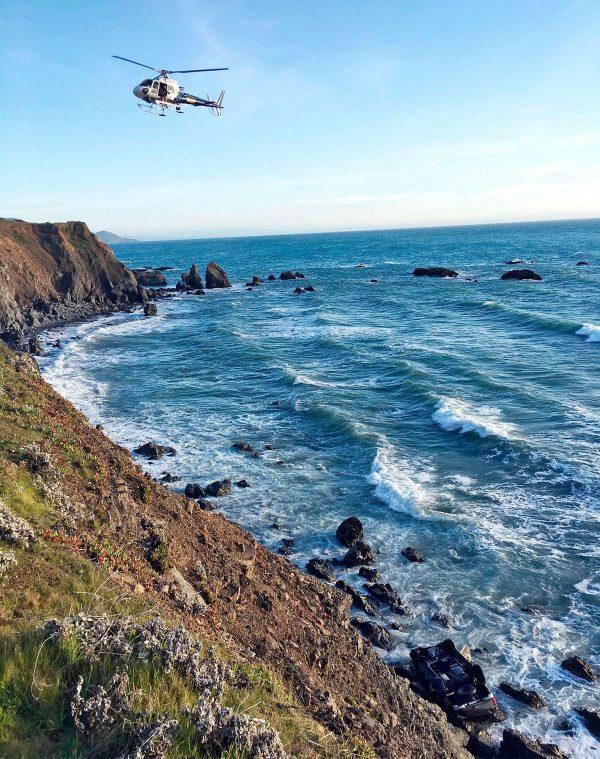 A helicopter hovers over steep coastal cliffs near Mendocino, Calif., where a vehicle, visible at lower right, plunged about 100 feet off a cliff along Highway 1, killing all passengers, on March 27, 2018. (California Highway Patrol via AP, File)