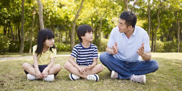 Asian father and two children sitting on grass having an interesting conversation, outdoors in a park. (Fotolia)