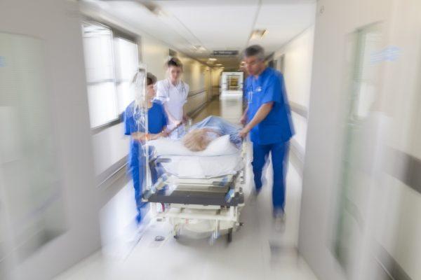 A motion blurred photograph of a patient on stretcher or gurney being pushed at speed through a hospital corridor by doctors & nurses to an emergency room. (Pxhere)
