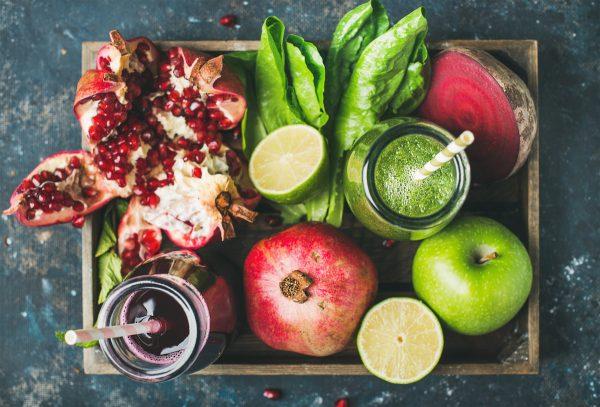 Green and purple fresh juices or smoothies with fruit, greens, vegetables in wooden tray over dark blue painted background, top view, selective focus. Detox, clean eating, vegetarian, vegan concept. (Shutterstock)