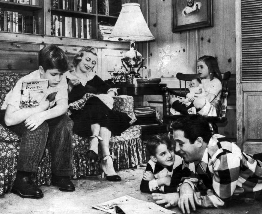Ronnie and Roselle Como sit on the sofa, Terri Como sits with her doll, David and Perry Como read on the floor at home in Long Island circa 1955 (©Wikimedia Commons | <a href="https://commons.wikimedia.org/wiki/File:Como_family_at_home_1955.jpg">Macfadden Publications</a>)