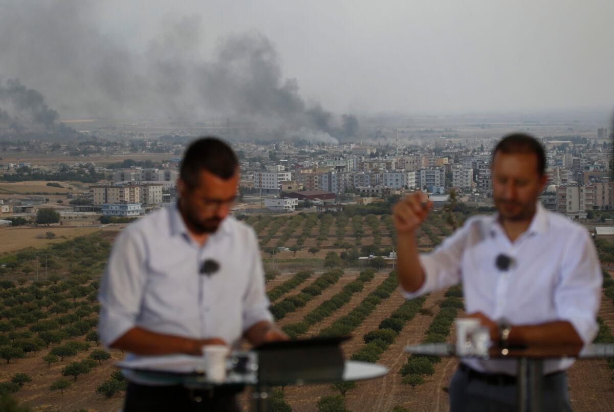 TV journalists talk during a live broadcast on a hilltop in Ceylanpinar, Sanliurfa province, southeastern Turkey, as in the background smoke billows from targets in Ras al-Ayn, Syria, during bombardment by Turkish forces on Oct. 16, 2019. (Lefteris Pitarakis/AP Photo)