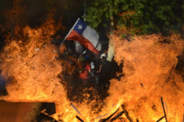 Seen through a burning street barricade, an anti-government demonstrator waves a Chilean flag in Santiago, Chile, on Oct. 28, 2019. (Matias Delacroix/AP Photo)