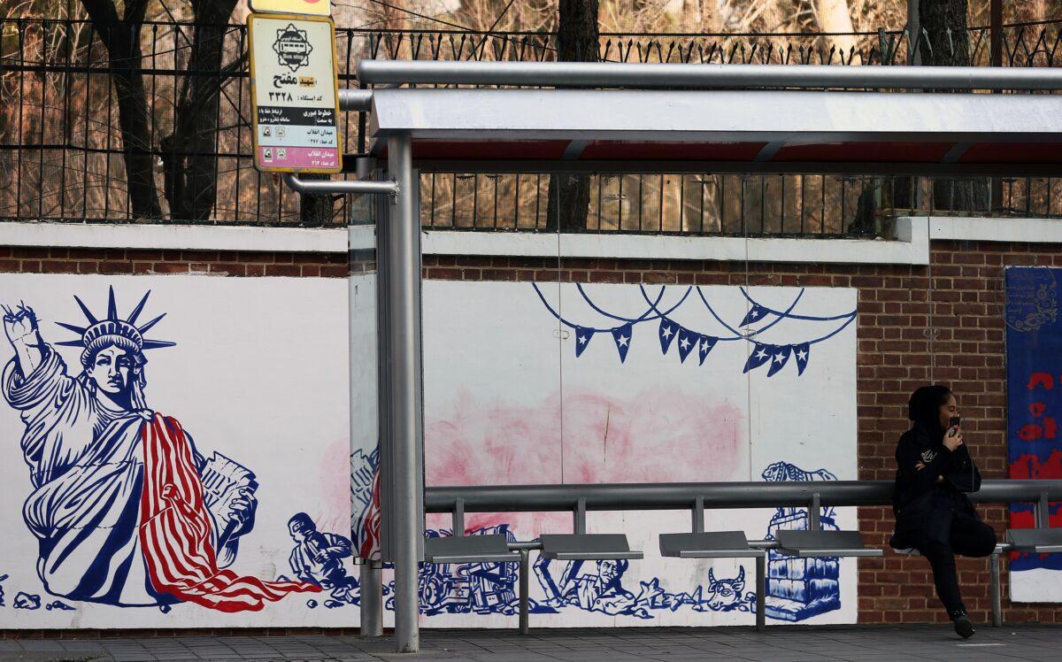 A woman sits at a bus stop in front of an anti-US mural on the former US embassy's wall in Tehran, Iran on Jan. 3, 2020. (Nazanin Tabatabaee/West Asia News Agency via Reuters)