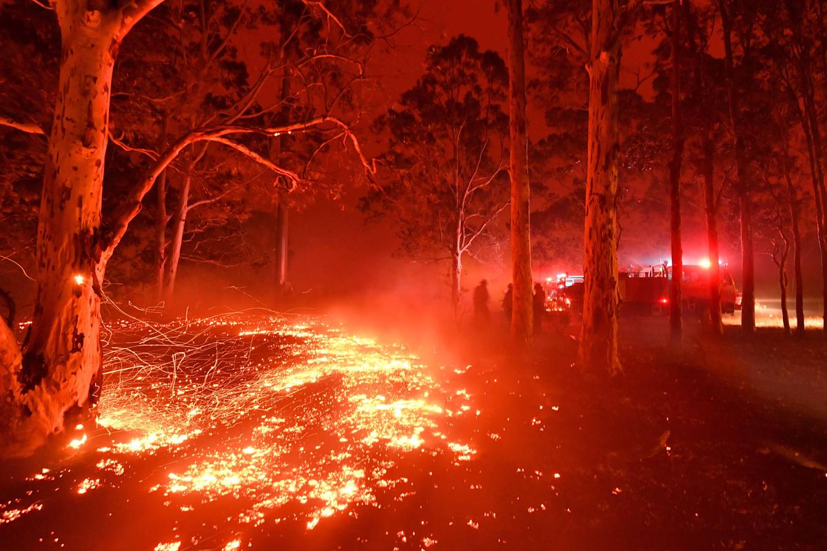 Burning embers cover the ground as firefighters (back R) battle against bushfires around the town of Nowra in the Australian state of New South Wales on Dec. 31, 2019. (©Getty Images | <a href="https://www.gettyimages.com/detail/news-photo/burning-embers-cover-the-ground-as-firefighters-battle-news-photo/1191093928?adppopup=true">SAEED KHAN</a>)